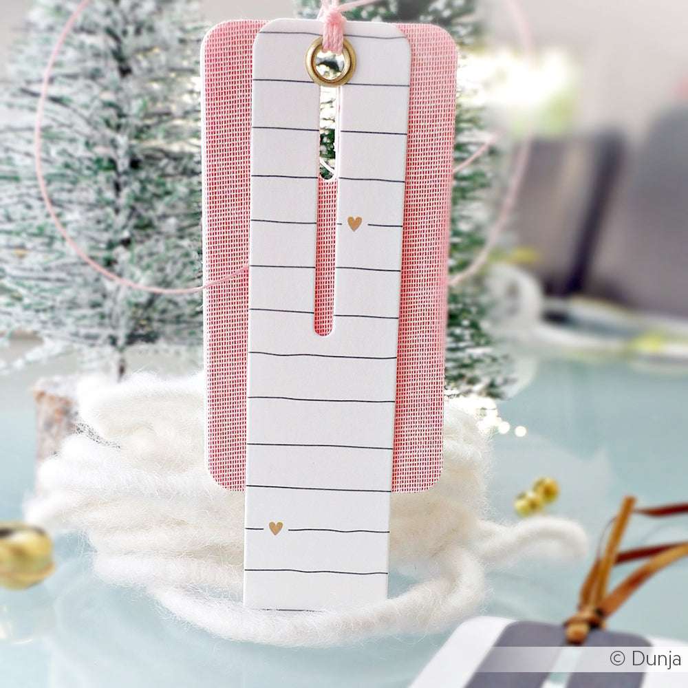 Zwei handgefertigte Lesezeichen von Alexandra Renke hängen vor einem verschneiten Mini-Weihnachtsbaum; eines ist weiß mit schwarzen Linien und goldenen Herzen.