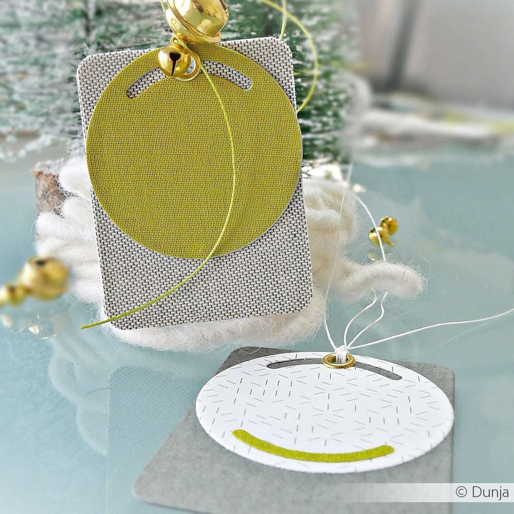 Zwei moderne Alexandra Renke Kombinations-Etikett-Geschenkanhänger mit geometrischen Motiven, goldener Kordel und Glockenakzenten sorgen für einen festlichen Touch auf einer spiegelnden Oberfläche.