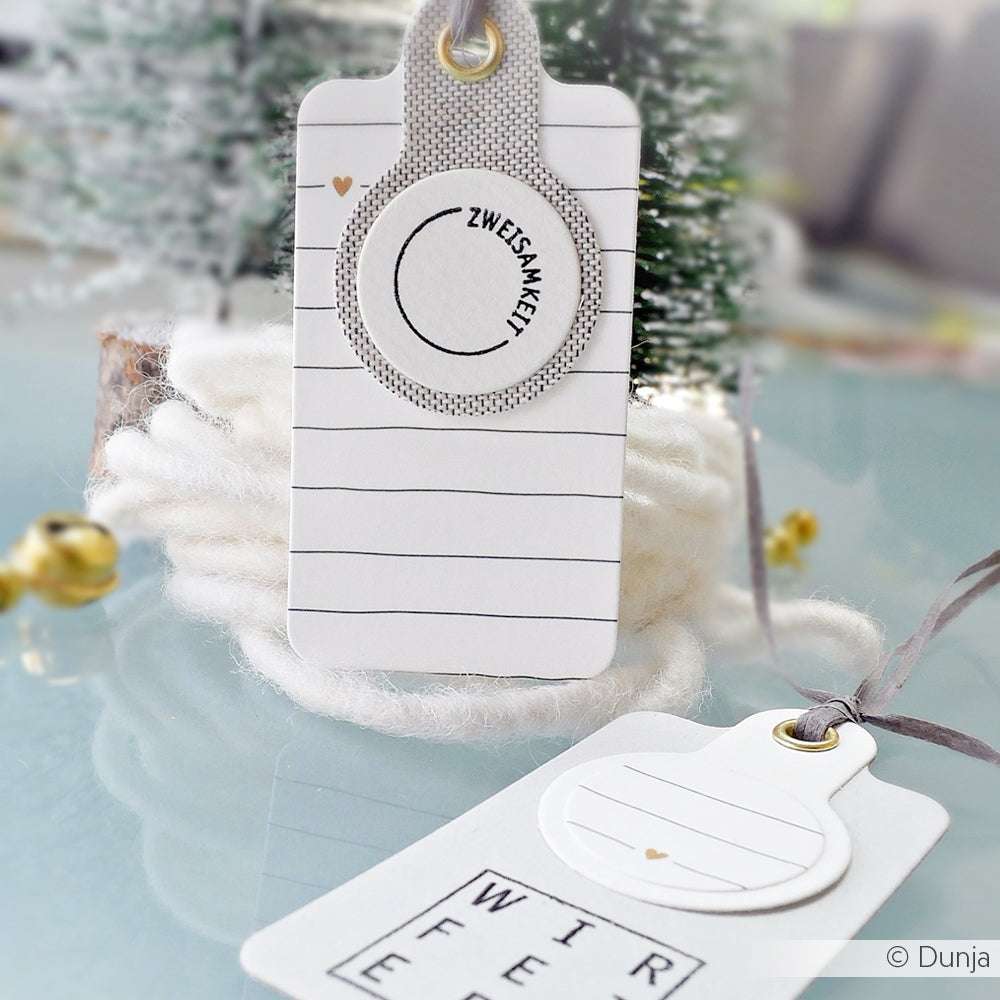 Zwei weiße, handgefertigte Geschenkanhänger von Alexandra Renke mit minimalistischem Design, goldenen Akzenten und deutschem Text auf hochwertigen Designpapieren, elegant arrangiert auf einer Glasfläche.