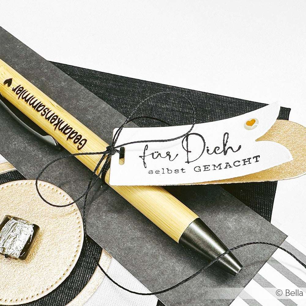 Ein mit schwarzer Schnur gebundener Stift mit der Aufschrift "für Dich selbst GEMACHT" ruht auf schwarz-goldenem Designpapier von Alexandra Renke.