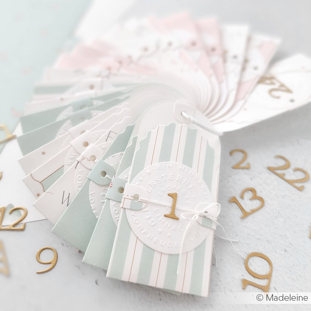 Gestreifte pastellfarbene Adventskalenderumschläge von Alexandra Renke, gefertigt aus eleganten Designpapieren und nummeriert mit goldenen Ziffern, stehen aufgefächert auf einem hellen Hintergrund.