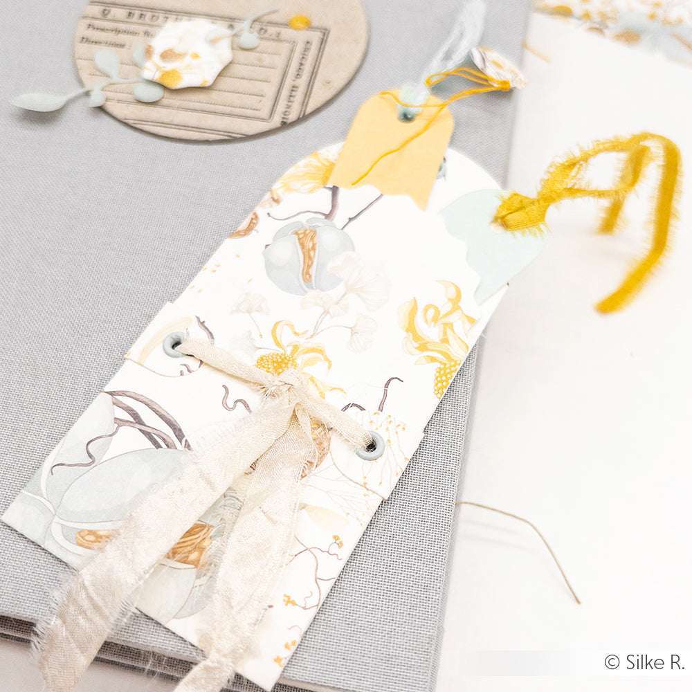 Dekorative florale Geschenkanhänger von Alexandra Renke, gefertigt aus Designpapier, veredelt mit Bändern und gelben Akzenten, dargestellt auf einem hellgrauen, strukturierten Hintergrund.