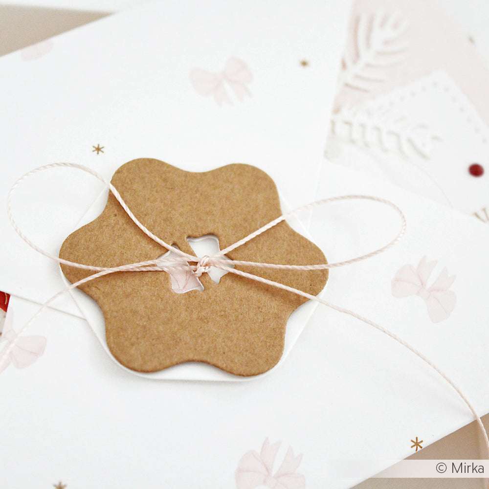 Ein blütenförmiger Knopf aus Kraftpapier, der mit einer Alexandra Renke Motivstanze gefertigt wurde, befestigt einen weißen Umschlag, der mit einer rosa Schleife verziert ist.