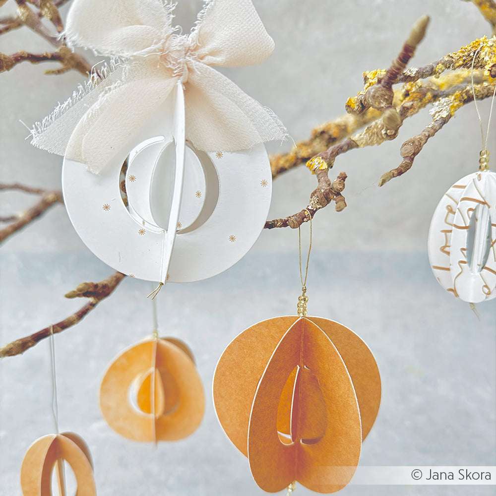 Alexandra Renke's Designpapiere und Motivstanze Kreis-Schnalle kreieren auffällige weiße und orangefarbene Papierornamente, die vor einem neutralen Hintergrund wunderschön an den Ästen hängen.