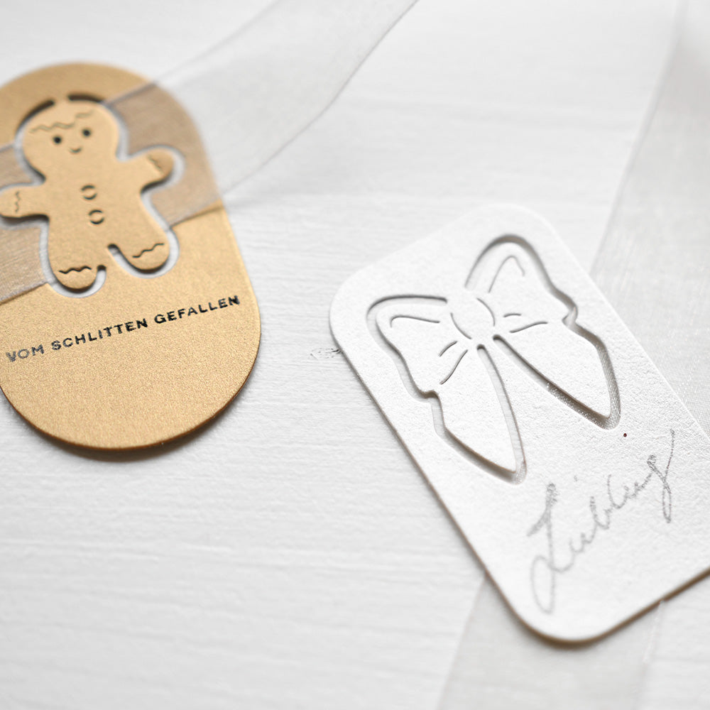 Goldfarbener Lebkuchenanhänger und weißer Schleifenanhänger mit deutschem Text auf hellem Hintergrund, hergestellt mit Alexandra Renke Designpapier und der Motivstanze Schleifen-Schnalle für elegante Details und durchsichtige Bandakzente.