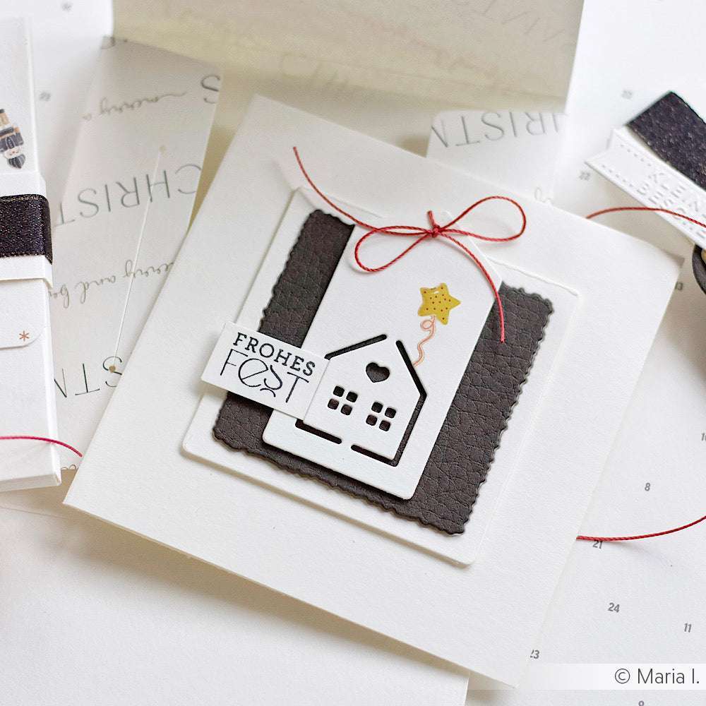 Handgefertigte Weihnachtskarte von Alexandra Renke mit einem mit Stahlstempeln gestanzten Hausmotiv in Weiß und Schwarz, das mit einer roten Schleife und dem Anhänger "Frohes Fest" versehen ist.