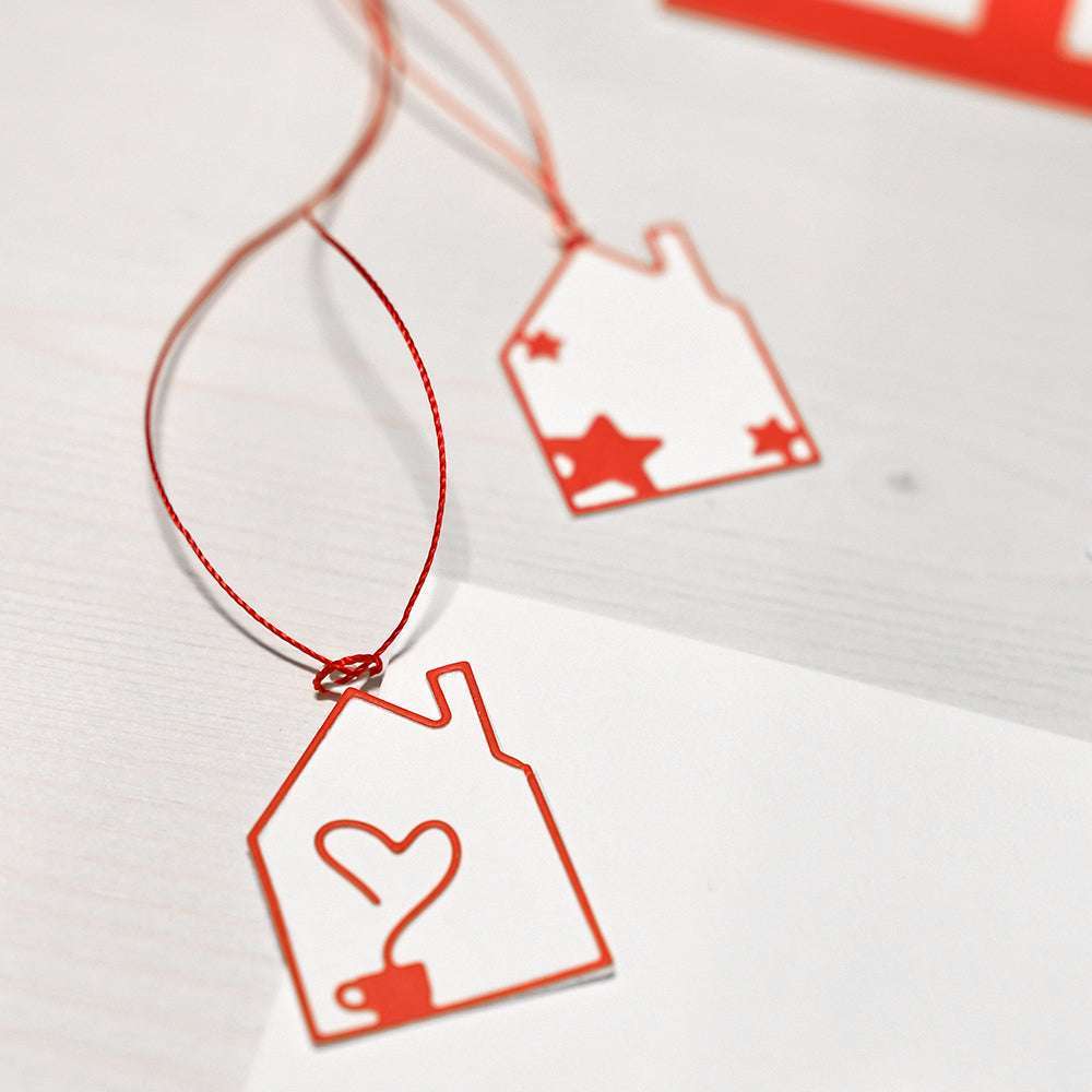 Zwei hausförmige rot-weiße Geschenkanhänger mit Herz- und Sternausschnitten, die mit einer Alexandra Renke Motivstanze Haus gefertigt wurden, liegen auf einer weißen Fläche.