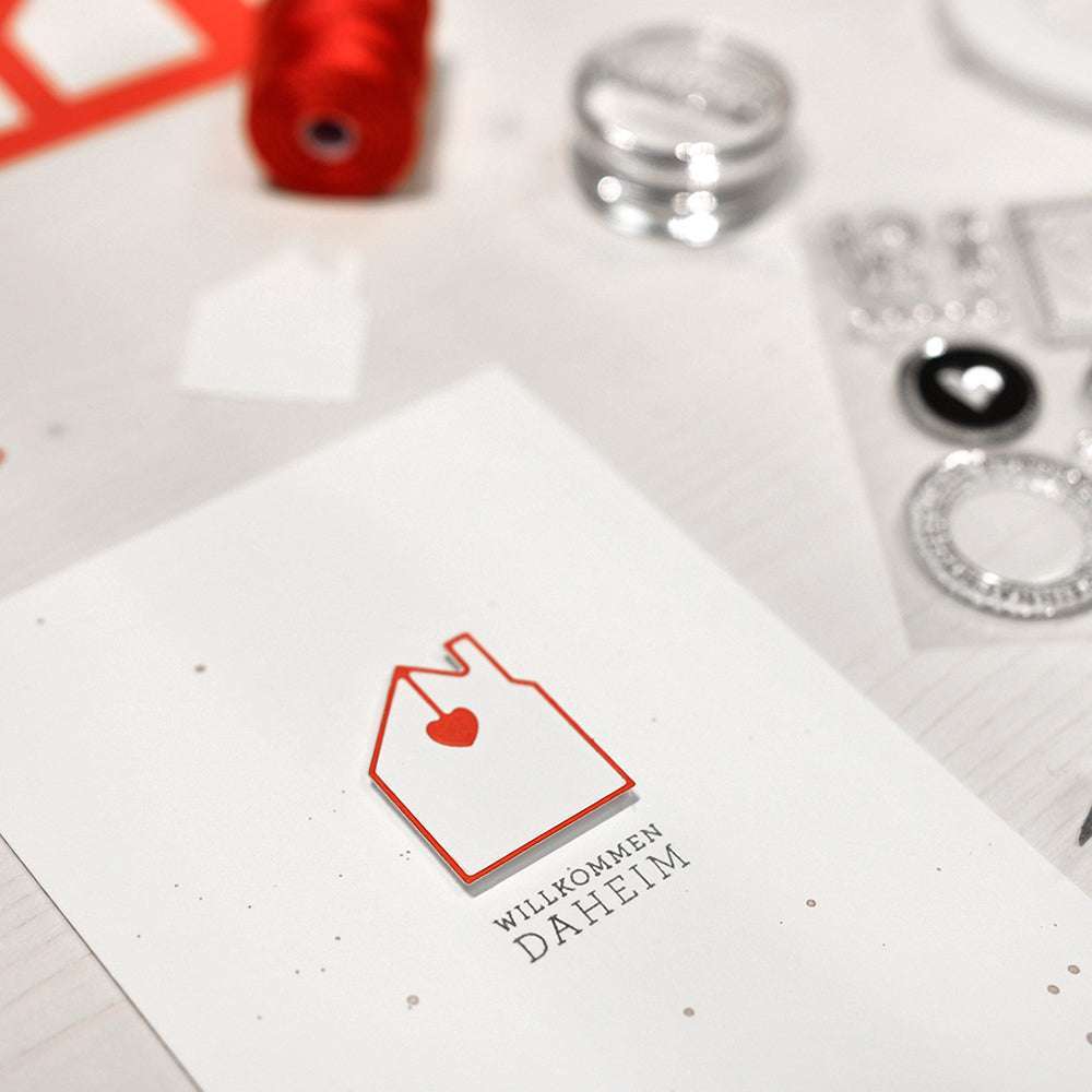 Auf einem hellen Tisch, umgeben von Bastelmaterial, liegt eine Karte mit einem roten Hausumriss mit Herz, hergestellt mit der Alexandra Renke Motivstanze Haus mit Herzanhänger-Rahmen, und dem Schriftzug "Willkommen Daheim".