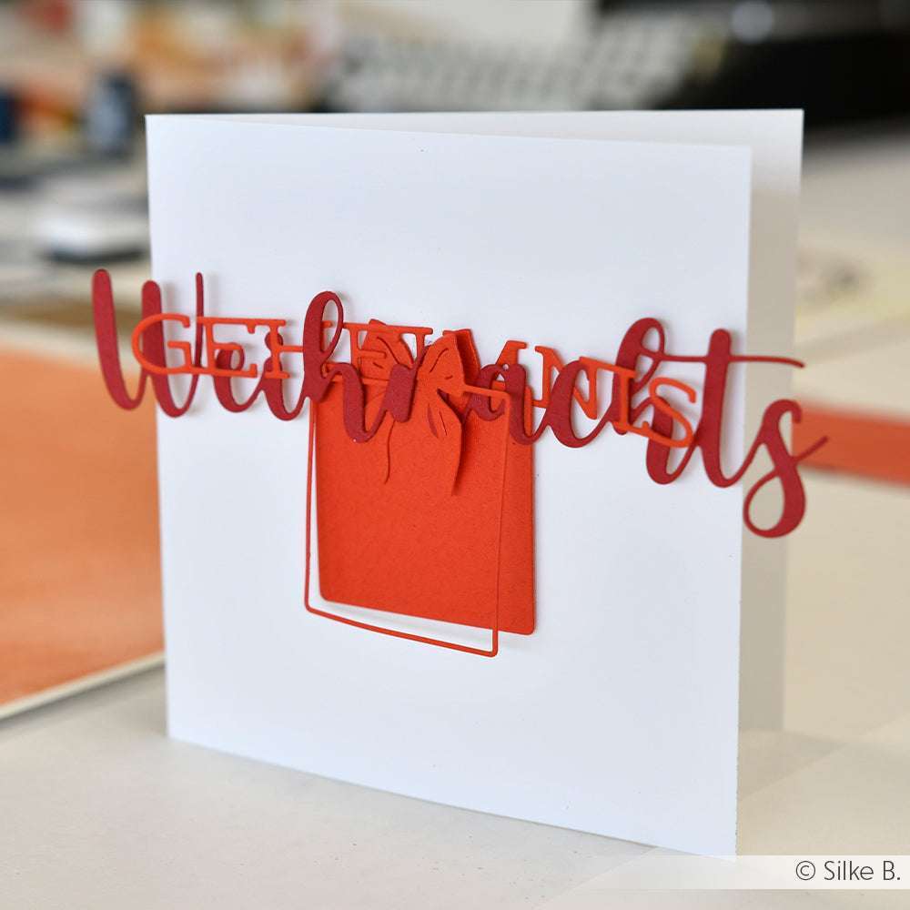 Weiße Grußkarte von Alexandra Renke mit roten Buchstaben und roter Geschenkbox auf der Vorderseite, hergestellt mit Stanzsystemen für Präzision und Designpapieren für eine hochwertige Verarbeitung.