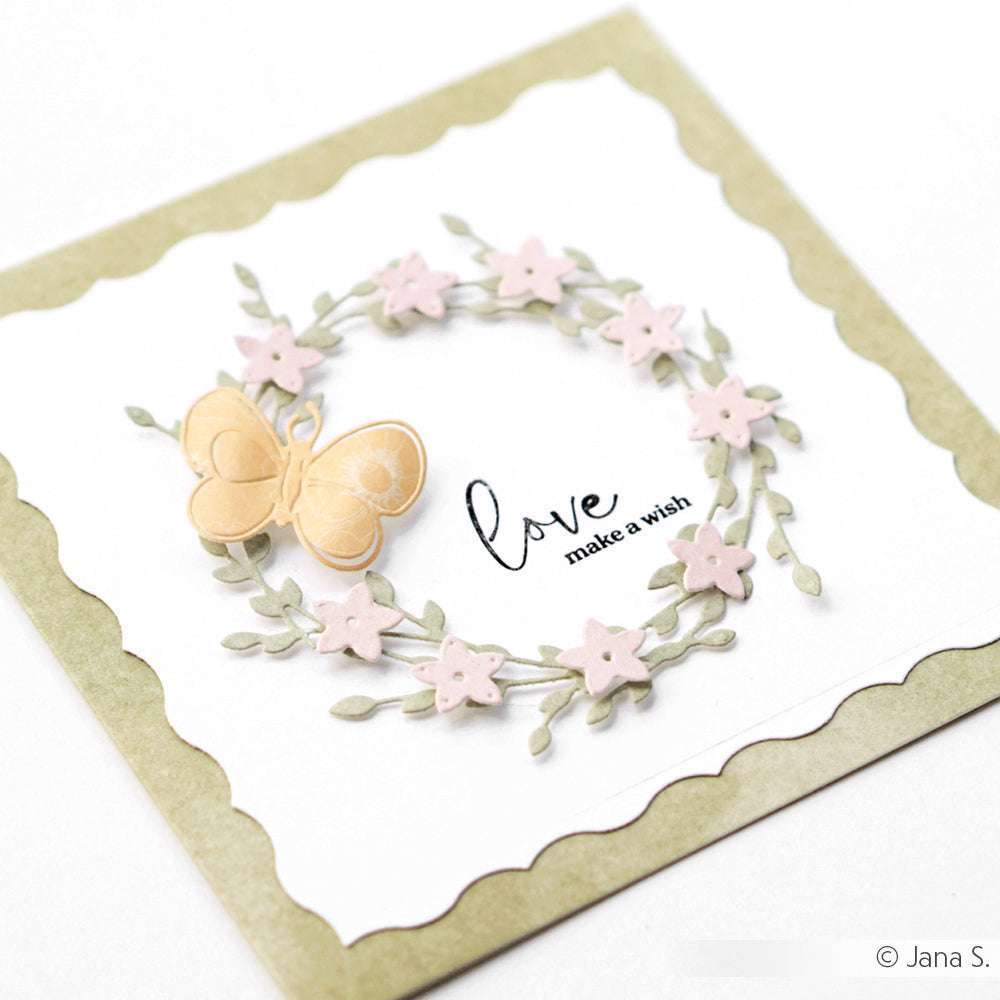 Handgefertigte Karte von Alexandra Renke mit einem Kranz aus rosafarbenen Blumen, einem beigen Schmetterling und dem Text "Love make a wish". Elegante Kartenaufleger und filigrane Stanzformen sorgen für zarte Details.