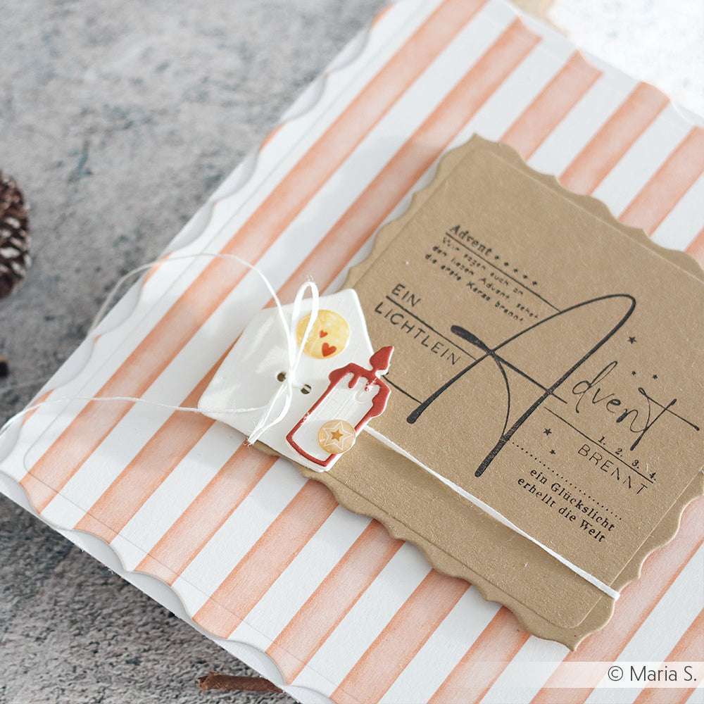 Ein in gestreiftes Papier eingepacktes Geschenk mit einer braunen Adventskarte und festlichen Anhängern, die mit einer Schnur verschnürt sind. Ein Alexandra Renke Ornamentrand Kartenaufleger sorgt für einen eleganten Touch.