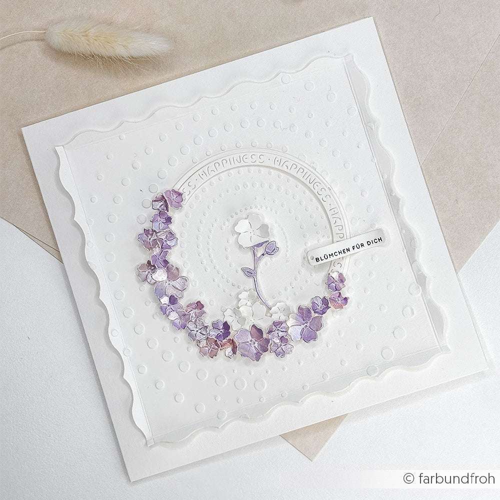 Eine zarte Karte von Alexandra Renke zeigt lila und weiße Papierblumen in einem Kranz, den deutschen Text "Blümchen für dich" und eine elegante Motivstanze Ornamentrand, die dem Kartenaufleger Charme verleiht.