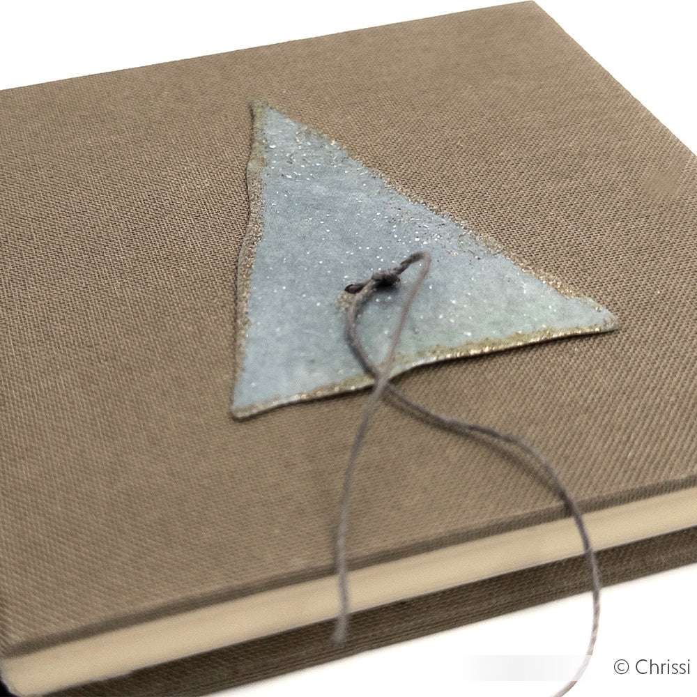 Ein braunes Hardcover-Buch von Alexandra Renke mit Designpapieren, einem strukturierten Dreieck, Motivstanze Dreieckige Knöpfe und Kordelverzierung auf dem Einband.