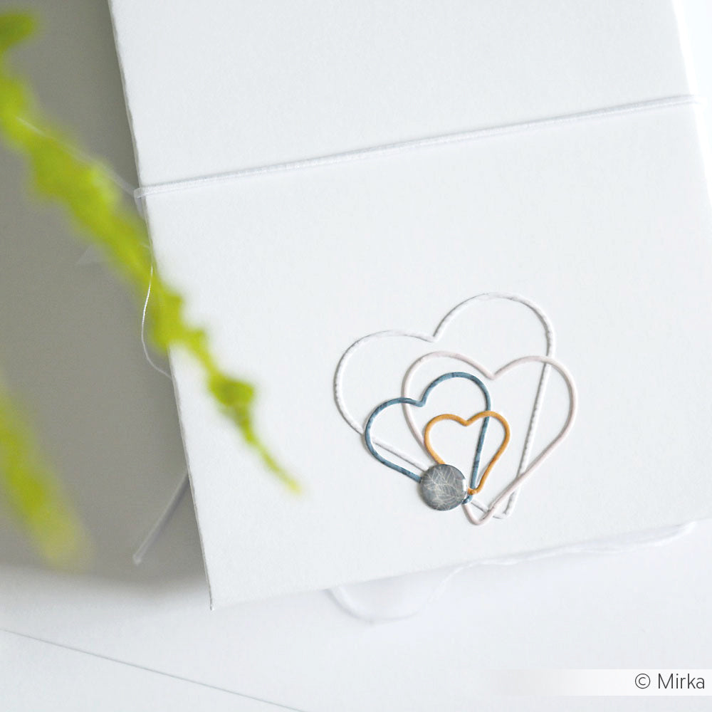 Weiße Karte mit drei gestickten, sich überlappenden Herzen in Blau, Gelb und Weiß, hergestellt mit den Motivstanze Herz Knöpfe von Alexandra Renke, neben einer grünen Pflanze.