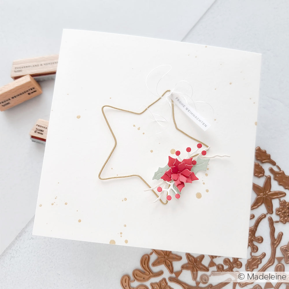 Eine weiße Grußkarte von Alexandra Renke mit einem goldenen Stern und roter Stechpalme, daneben Stempel, Designpapier und eine Motivstanze aus Metall.