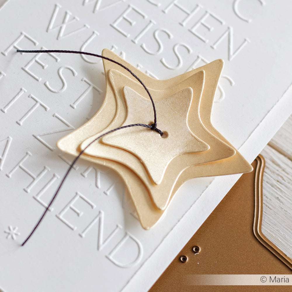 Drei übereinander liegende goldene Stern-Ornamente, die mit Alexandra Renke Motivstanze Stern Knöpfe gefertigt wurden, hängen an einer schwarzen Schnur auf einem weißen Hintergrund aus geprägter Karte.