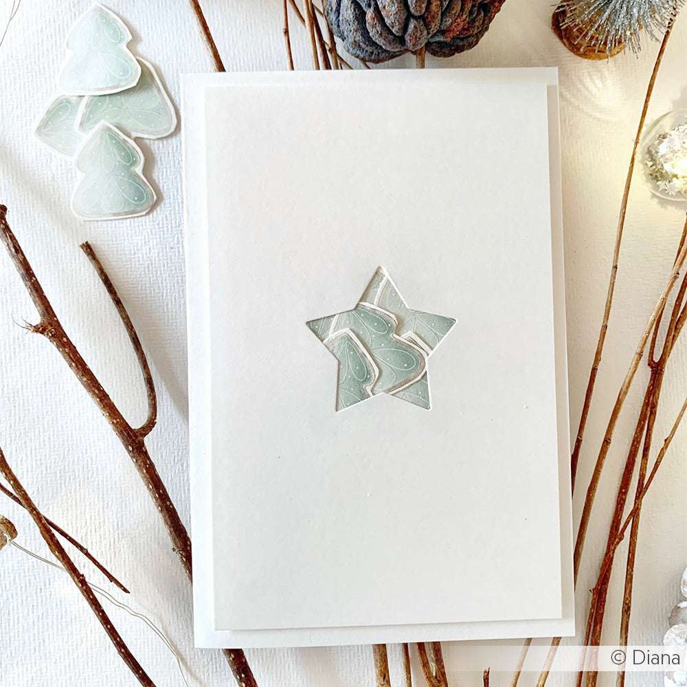 Weiße Karte mit einem marmorierten Sternausschnitt, der mit der Alexandra Renke Motivstanze Stern Etikett hergestellt wurde und auf einer weißen Fläche, umgeben von Zweigen und herzförmigen Objekten, präsentiert wird.