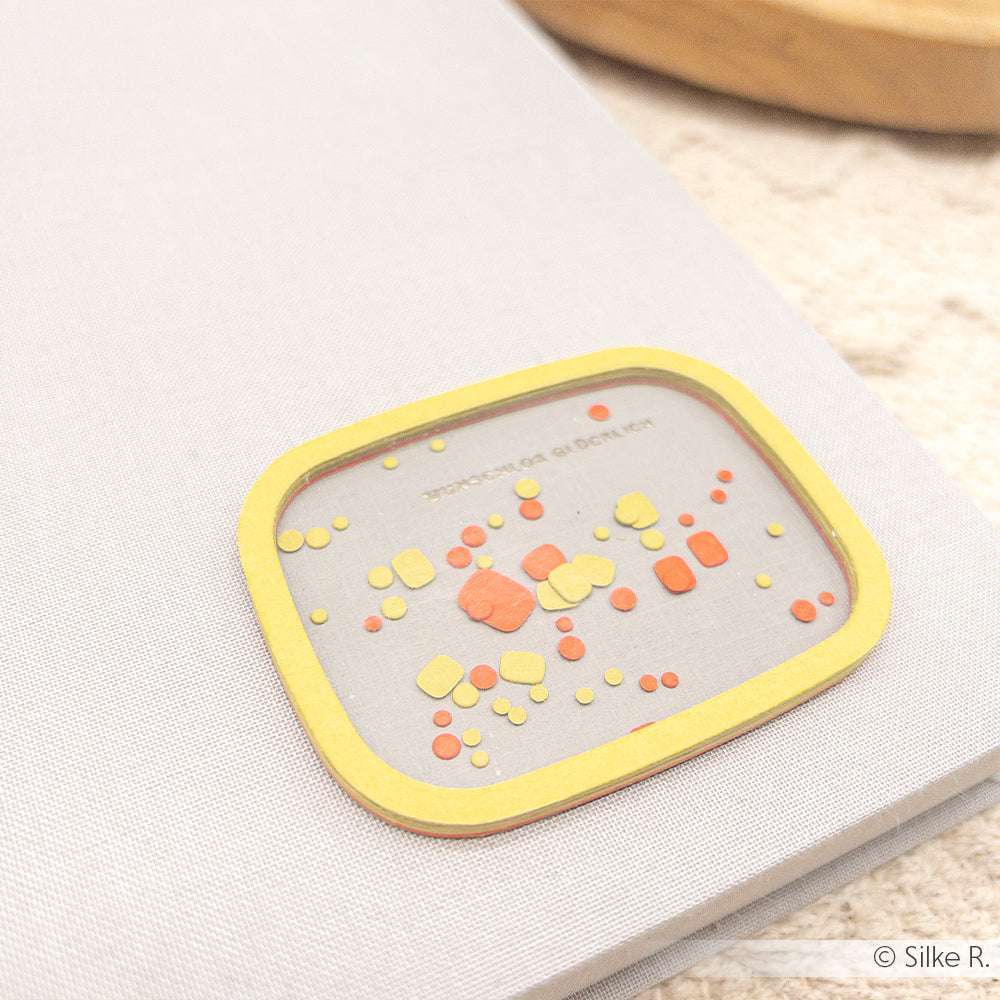 Ein kleiner rechteckiger Alexandra Renke PAPPcut®ies Aufkleber mit gelben und orangen Punkten auf einem grauen Notizbuch.