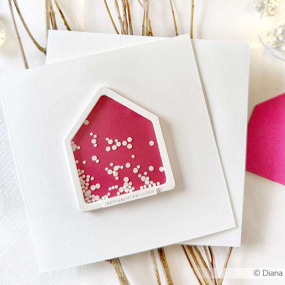 Eine rosafarbene Karte in Form eines Hauses, die mit der Alexandra Renke Haus-Rahmen-Stanzform hergestellt wurde, ist mit weißem Konfetti verziert und steht auf makellos weißem, quadratischem Papier.