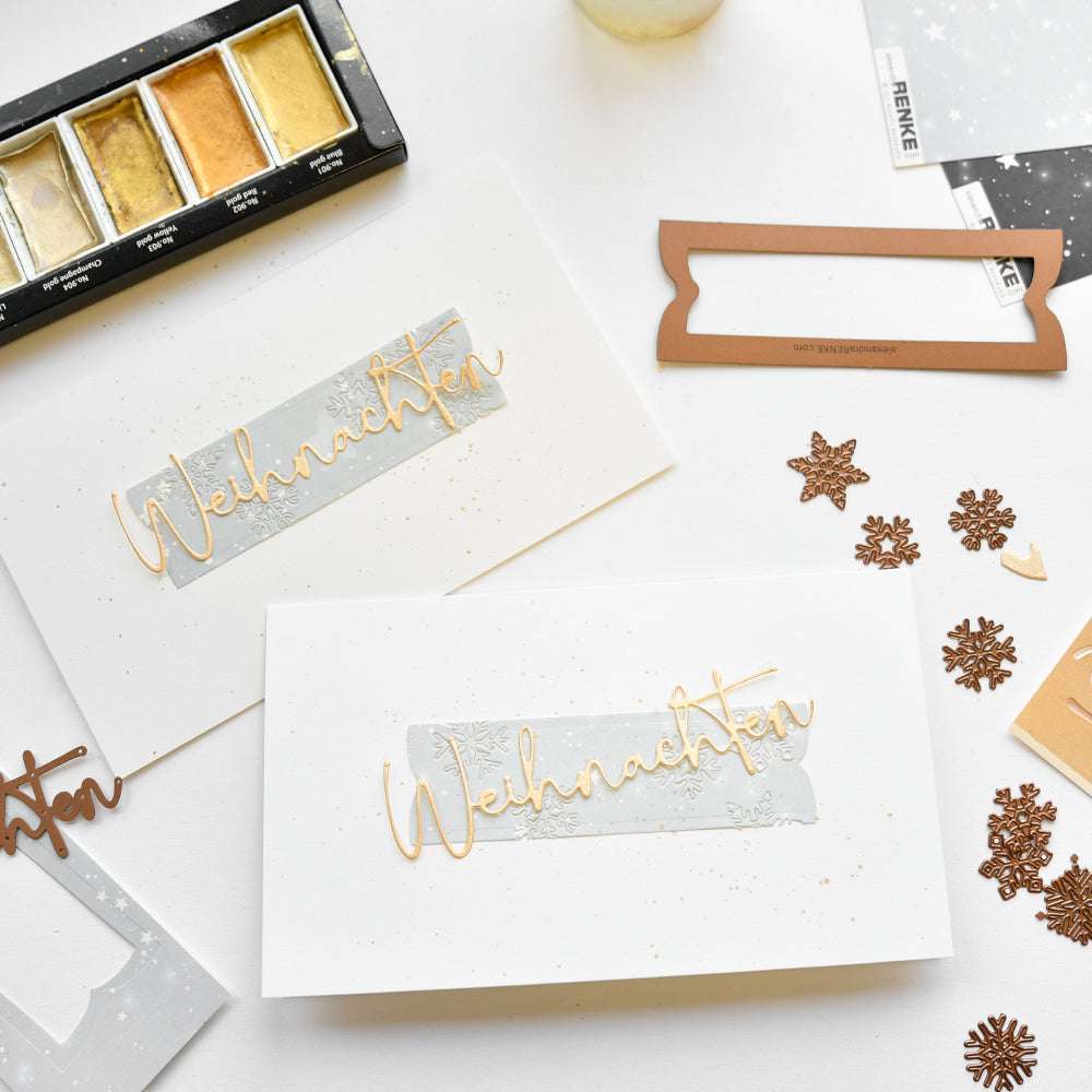 Zwei handgefertigte Weihnachtskarten mit "Weihnachten" in goldener Schrift, hergestellt mit Alexandra Renke Designpapieren und einer Motivstanze Rechteckiger Kartenaufleger, umgeben von Bastelmaterial und Schneeflockenausschnitten.