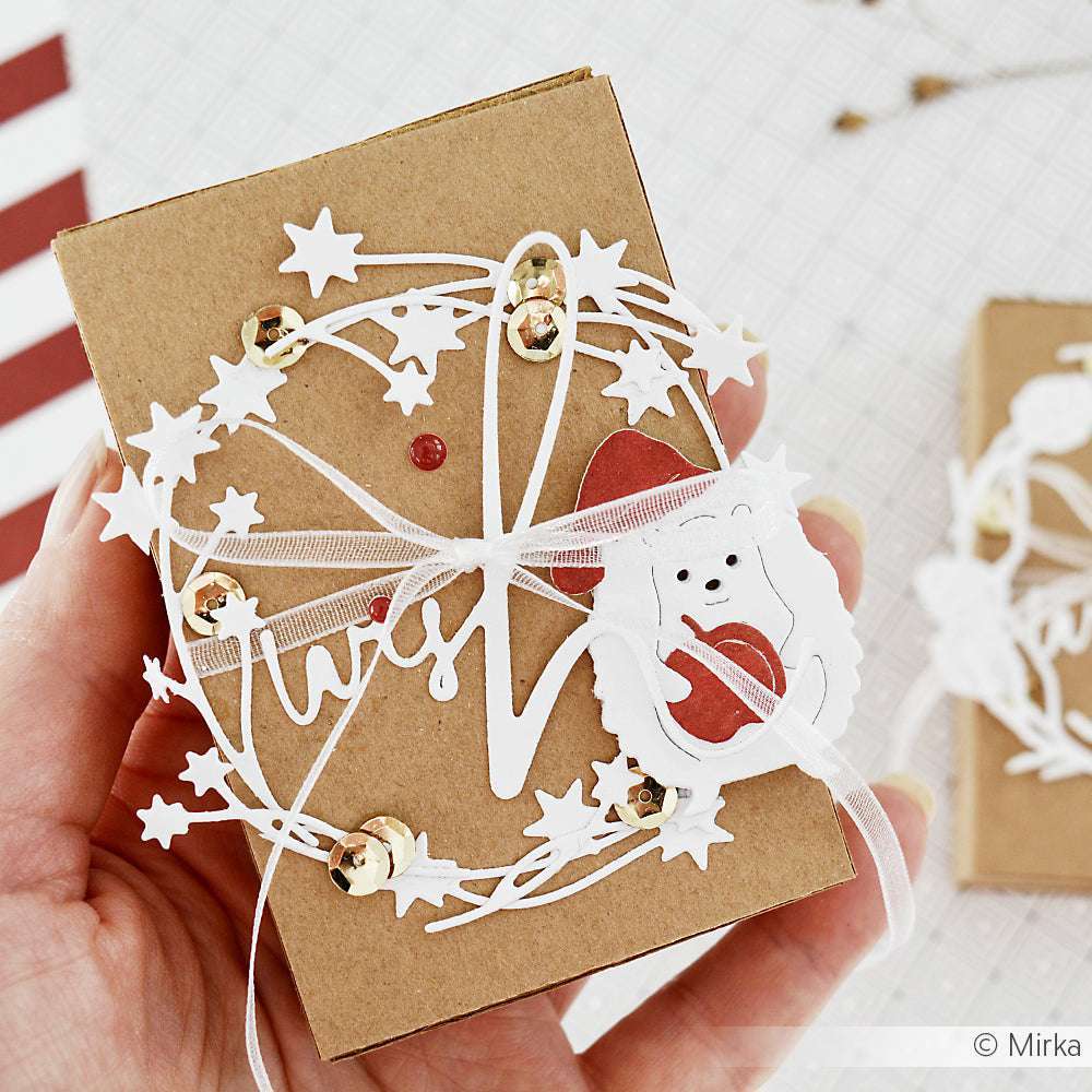 In der Hand hält er eine dekorierte braune Geschenkbox mit weißen Ornamenten, Sternen, Pailletten und einem ausgeschnittenen Bären, der ein rotes Herz hält - die perfekte Inspiration für Alexandra Renke Stanzen für Papierbasteln und kreative DieCut Systeme.