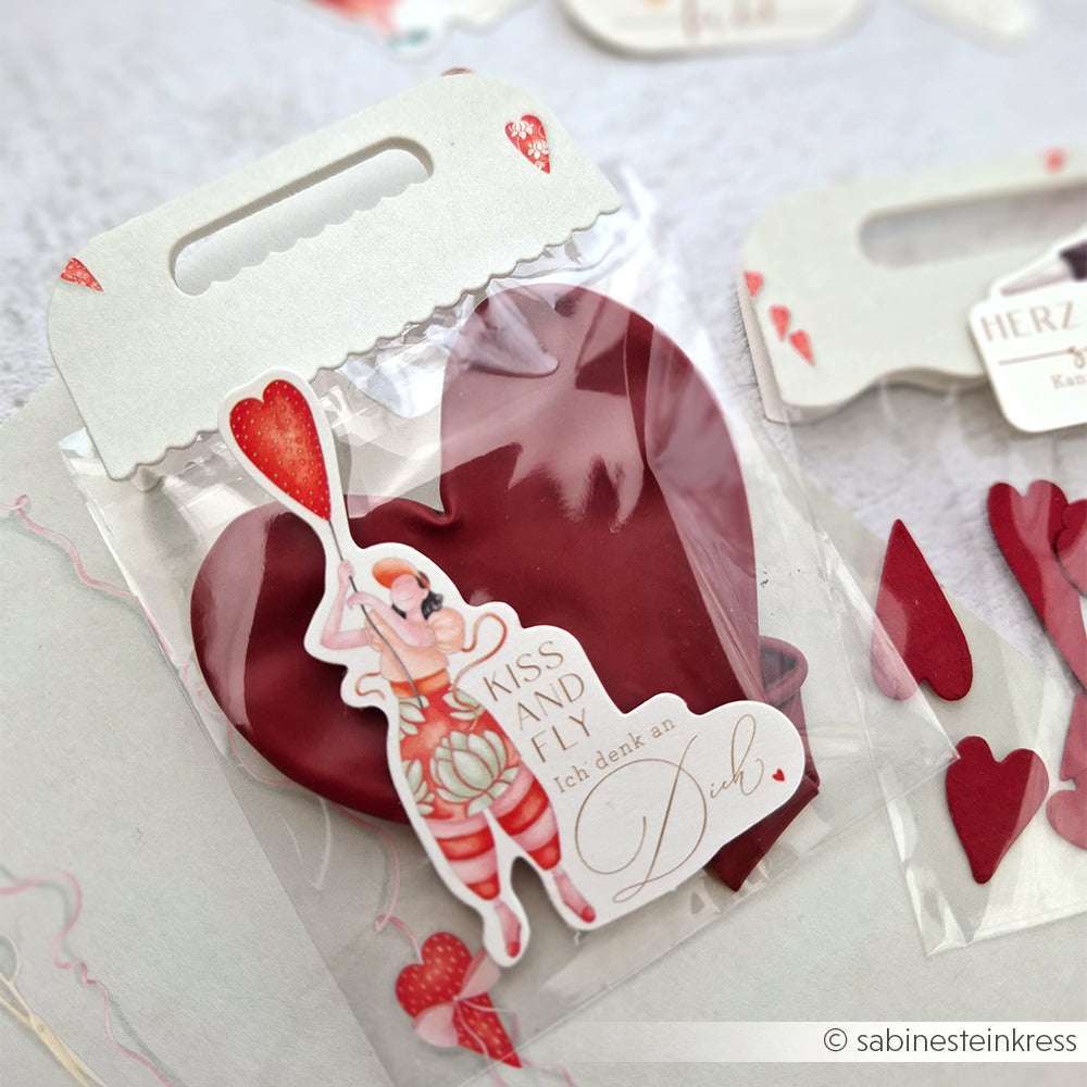 Eine rote Herzschokolade von Alexandra Renke in einem durchsichtigen Beutel mit einem Cartoon-Anhänger mit der Aufschrift "Kiss and Fly!" und einem schicken Motivstanze-Tütengriff für eine besondere Note.