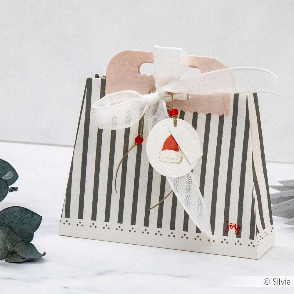 Alexandra Renke gestreifte Geschenktüte mit weißem Band, roten Perlen und Weihnachtsmannmütze aus festlichen Designpapieren, Henkel geformt mit der Motivstanze Tütengriff, ausgestellt auf einer weißen Fläche.