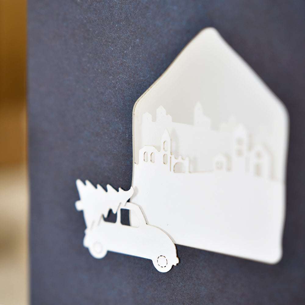 Weißer Papierausschnitt eines Lastwagens, der einen Baum trägt, mit Haus- und Stadtsilhouetten auf dunklem Hintergrund - hergestellt mit Alexandra Renke Motivstanze Mini Landschaft und hochwertigen Designpapieren für elegante Details.