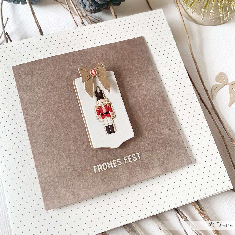 Eine Weihnachtskarte von Alexandra Renke, erstellt mit Designpapieren, zeigt einen Nussknacker-Dekor, eine goldene Schleife, erstellt mit Motivstanze Schicht Schleife, und den Text Frohes Fest" auf einem braunen quadratischen Hintergrund.