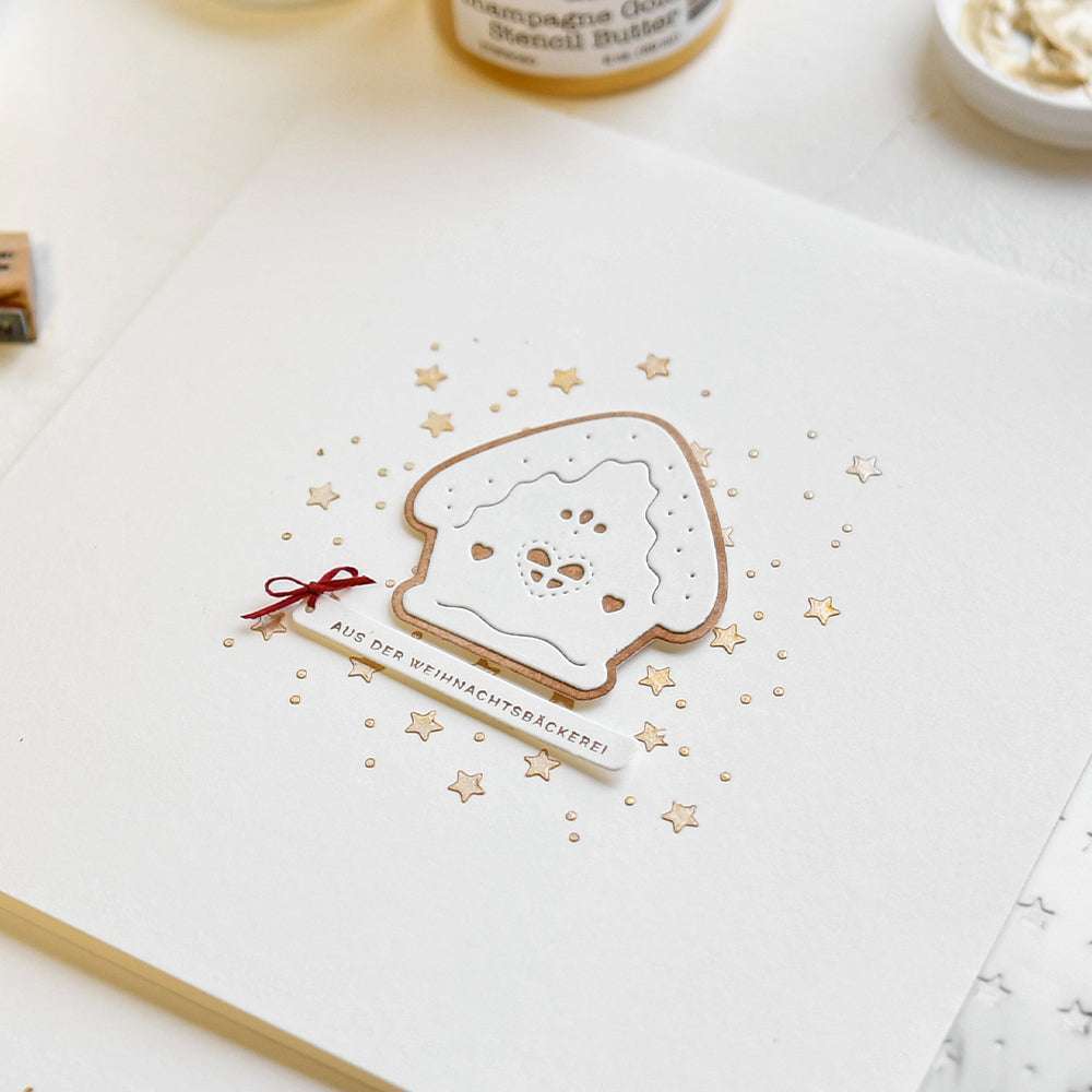 Eine handgemachte Weihnachtskarte von Alexandra Renke mit einem ausgestanzten Lebkuchenhaus, goldenen Sternen und einer kleinen roten Schleife auf weißem Papier.