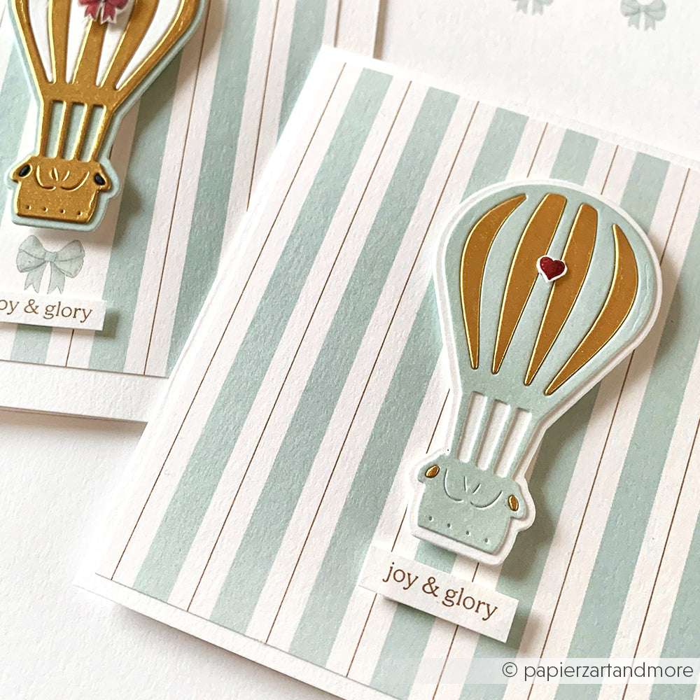 Grußkarte von Alexandra Renke mit einem goldenen und mintfarbenen Heißluftballon, der mit einer Heißluftballon-Stanzform hergestellt wurde und mit einem joy & glory-Etikett auf einem gestreiften Hintergrund verziert ist.