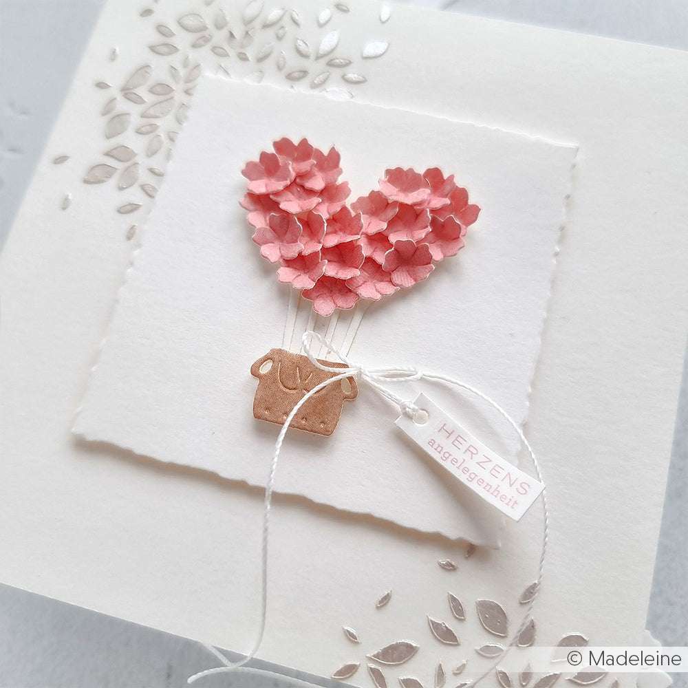 Handgemachte Karte von Alexandra Renke mit rosafarbenen Papierblumen in einem herzförmigen Topf, die mit Designpapier gestaltet und mit einem kleinen Anhänger versehen sind.