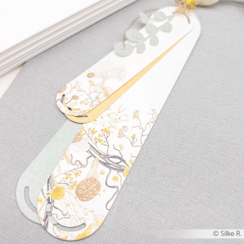 Zwei elegante, floral gemusterte Lesezeichen von Alexandra Renke, gefertigt aus hochwertigen Designpapieren mit gelben, goldenen und grauen Akzenten, liegen auf hellgrauem Untergrund, der mit der Stanzsysteme Motivstanze Elegantes Etiketten Duo 4 veredelt wurde.
