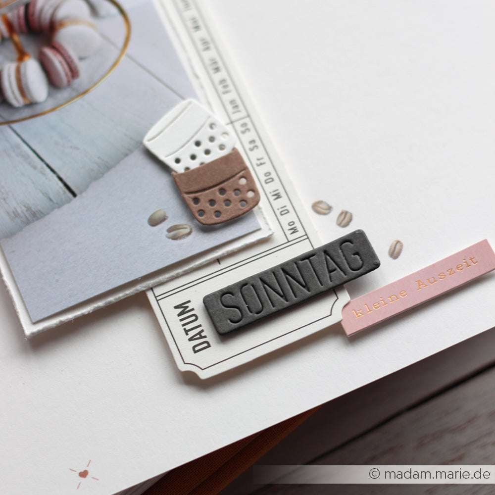 Scrapbook-Seite von Alexandra Renke mit einem Foto, Ausschnitten zum Thema Kaffee, dem Etikett "SONNTAG" aus der Motivstanze Wochentage Schildchen, dekorativen Anhängern und Designpapieren auf einem sauberen weißen Hintergrund.