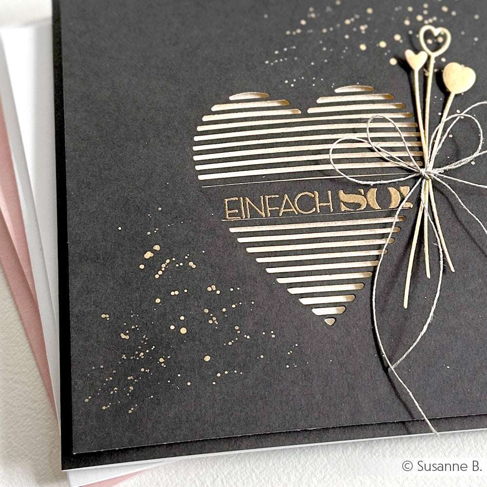 Schwarze Grußkarte mit einem ausgeschnittenen, gestreiften Herz aus der Alexandra Renke Motivstanze Lamellen Herz, akzentuiert durch goldene Spritzer und eine Kordel mit kleinen Herzen - ideal für schicke, handgemachte Grüße.