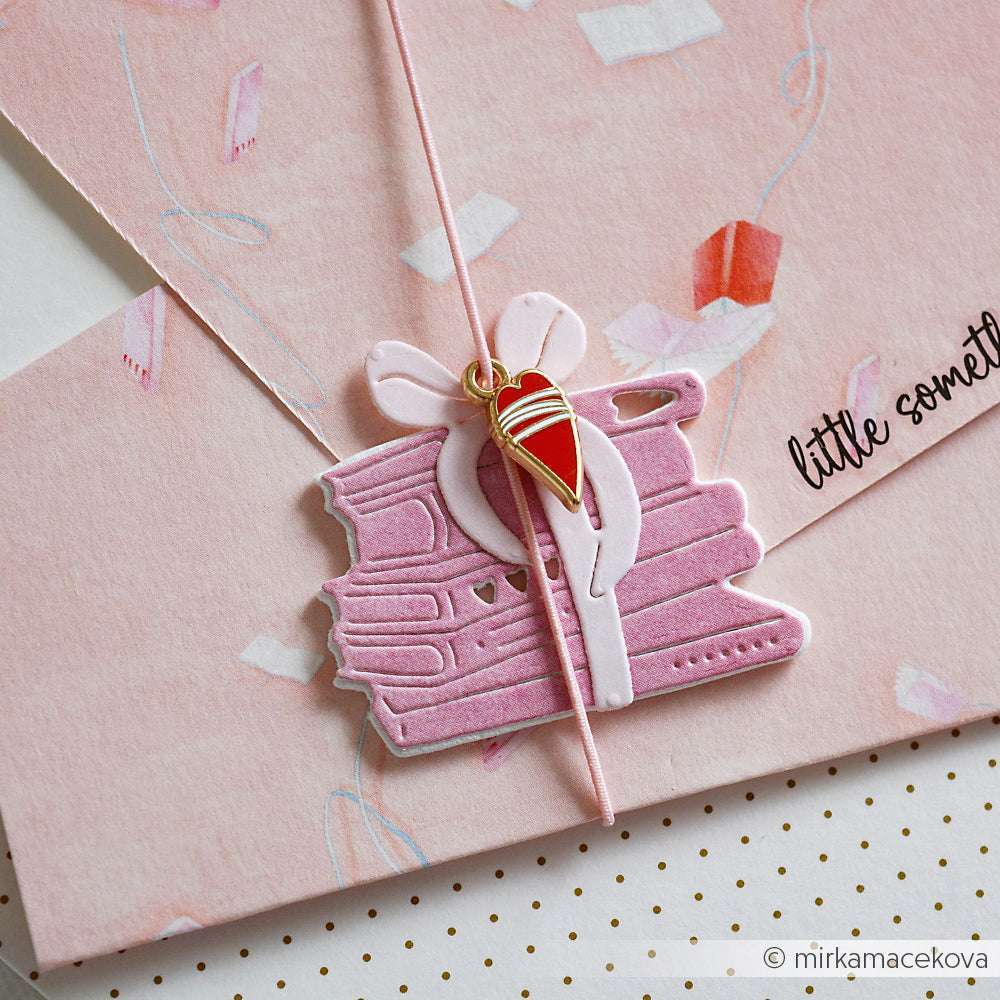 Rosafarbener Umschlag mit gestapelten Büchern und rotem Herzanhänger, gebunden mit einer rosafarbenen Kordel - perfekt zum Verschenken, vor allem in Kombination mit Ihren Lieblingskreationen von Alexandra Renke.