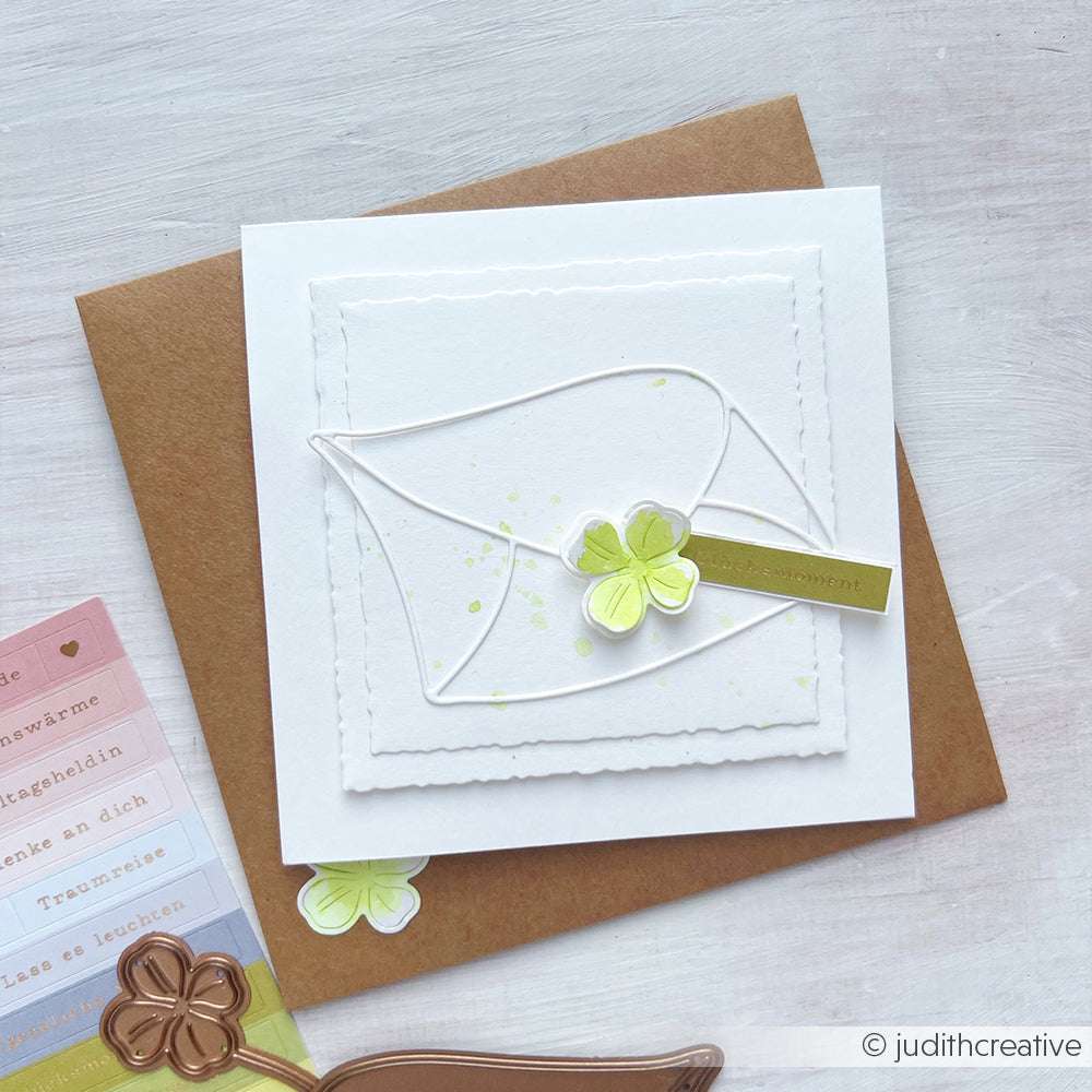 Quadratische Karte mit grünem Kleeblatt, weißem Umschlag und Gruß auf braunem Umschlag, gestaltet mit Alexandra Renke Motivstanze und PAPPcut®ies, mit Bastelmaterial in der Nähe.