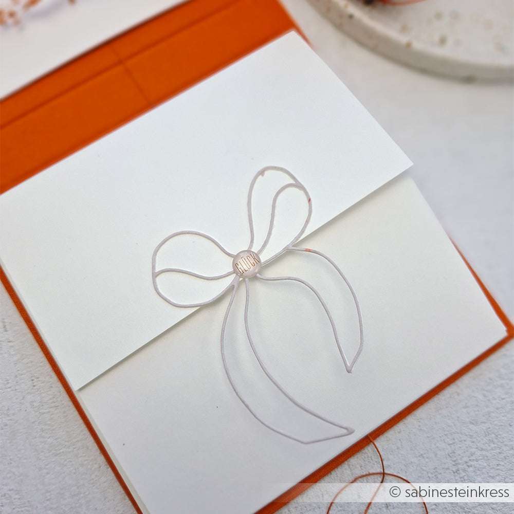 Weißer Umschlag mit minimalistischer Drahtschleife, gefertigt mit der Motivstanze Schleife von Alexandra Renke, auf orangefarbenem Hintergrund.