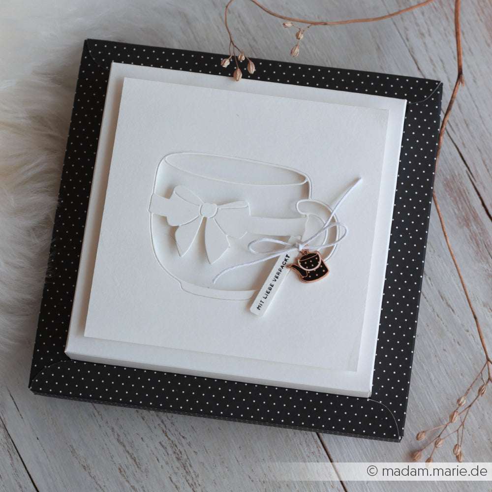 Eine weiße Grußkarte mit einer Teetasse und einer Schleife, die mit der Motivstanze Große Schleifentasse von Alexandra Renke gestaltet wurde, ruht auf einer schwarz gepunkteten Schachtel auf einer Holzoberfläche.