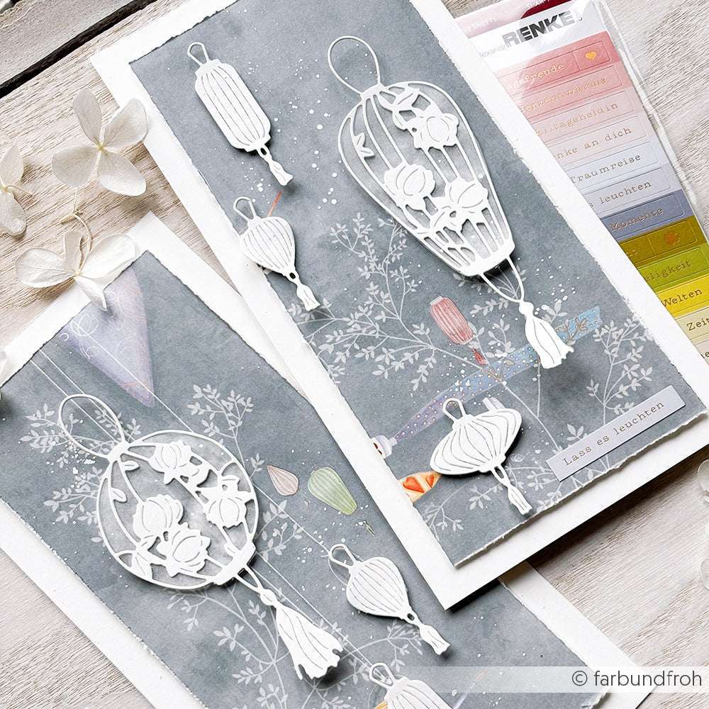 Handgemachte Karten von Alexandra Renke mit verschlungenen weißen Papierlaternen auf blau-grauem Blumenhintergrund, akzentuiert mit farbigen Laternen in der Ecke.