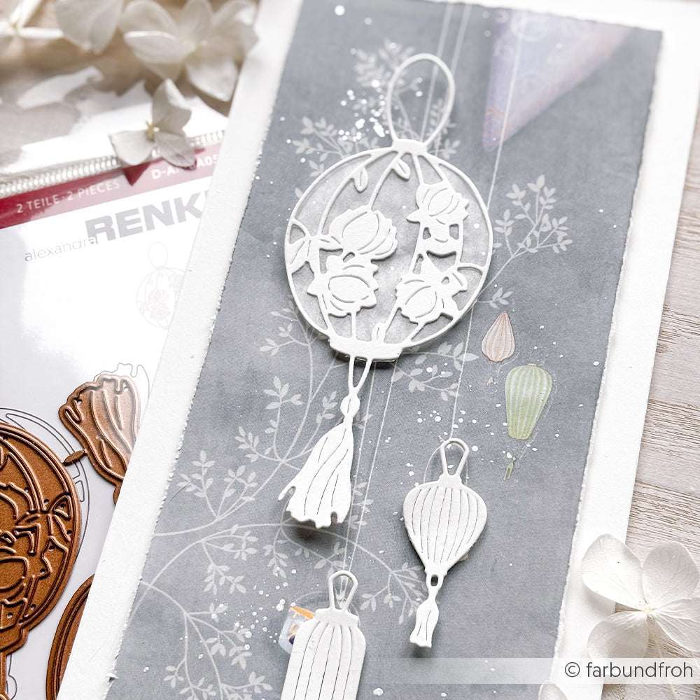 Weiße Blumen und Laternen, die mit Alexandra Renke Deep Edge Motivstanzen und PAPPcut®ies ausgeschnitten wurden, werden vor einem grauen, botanischen Hintergrund bei sanfter Beleuchtung präsentiert.