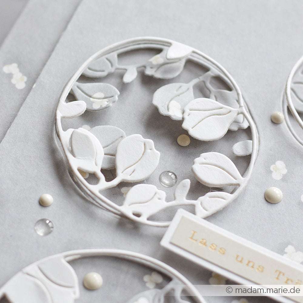 Nahaufnahme eines zarten, weißen, floralen Scherenschnittmusters auf hellgrauem Hintergrund mit Zierperlen, hergestellt mit Alexandra Renke Deep Edge Motivstanzen für gestochen scharfe Details.