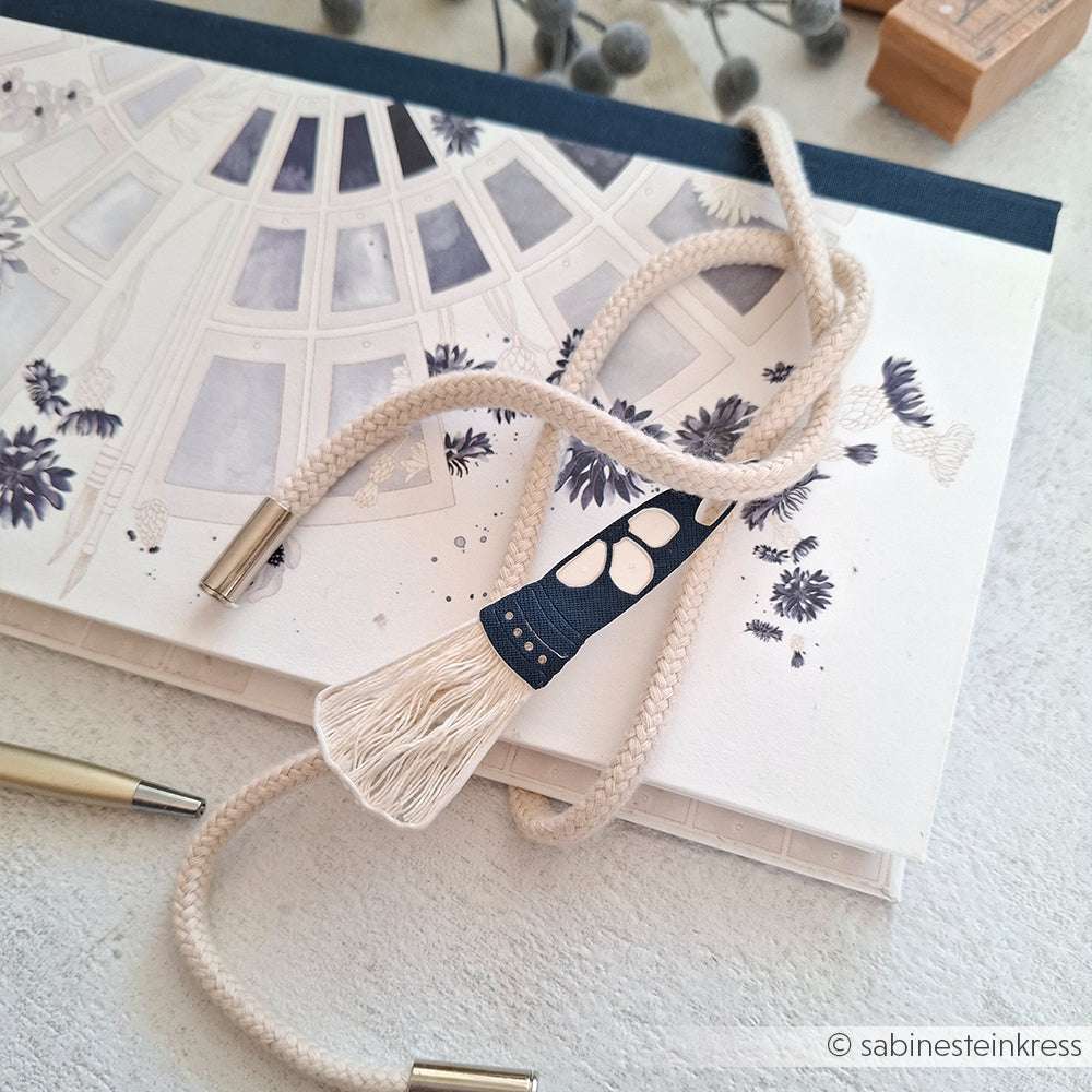 Eine geflochtene Kordel mit geflochtenen Enden ruht auf einem botanischen Skizzenbuch, das mit zarten Formen aus Alexandra Renke Deep Edge Motivstanzen verziert ist.