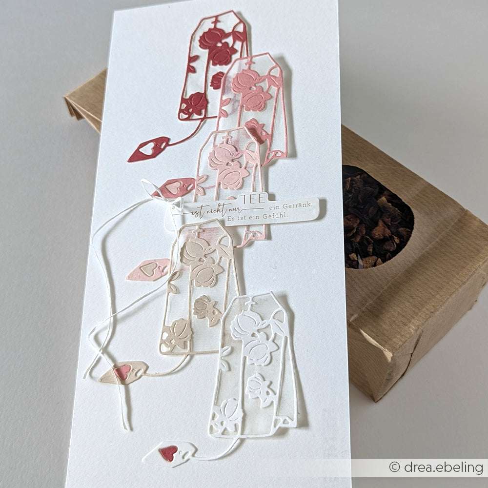 Handgefertigte Karte mit geschichteten, ausgeschnittenen Teebeuteldesigns aus der Motivstanze Großer blumiger Teebeutel von Alexandra Renke in Rosa und Weiß, platziert auf einer Schachtel mit losen Teeblättern.