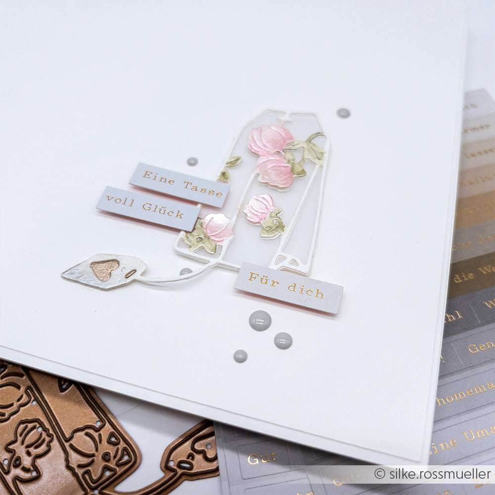 Eine handgefertigte Karte von Alexandra Renke mit einer Teetasse, rosafarbenen Blumen und dem Text "Eine Tasse voll Glück Für dich", gefertigt mit einer Motivstanze großer blumiger Teebeutel für zusätzlichen Charme.
