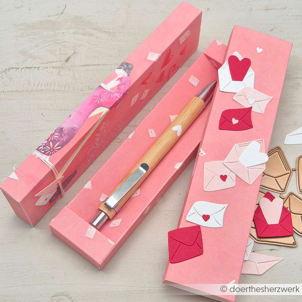 Rosa Geschenkbox von Alexandra Renke mit Bambusstift, verziert mit Deep Edge Motivstanzen und PAPPcut®ies Papierherzen und Briefumschlagausschnitten, ausgestellt auf einer hellen Holzoberfläche.