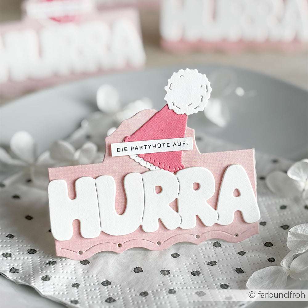 Rosa-weiße Karte mit "HURRA" und einem Partyhut, hergestellt mit der Alexandra Renke Motivstanze Kleine Partyhüte, präsentiert auf einer gepunkteten Serviette mit weißen Blütenblättern.