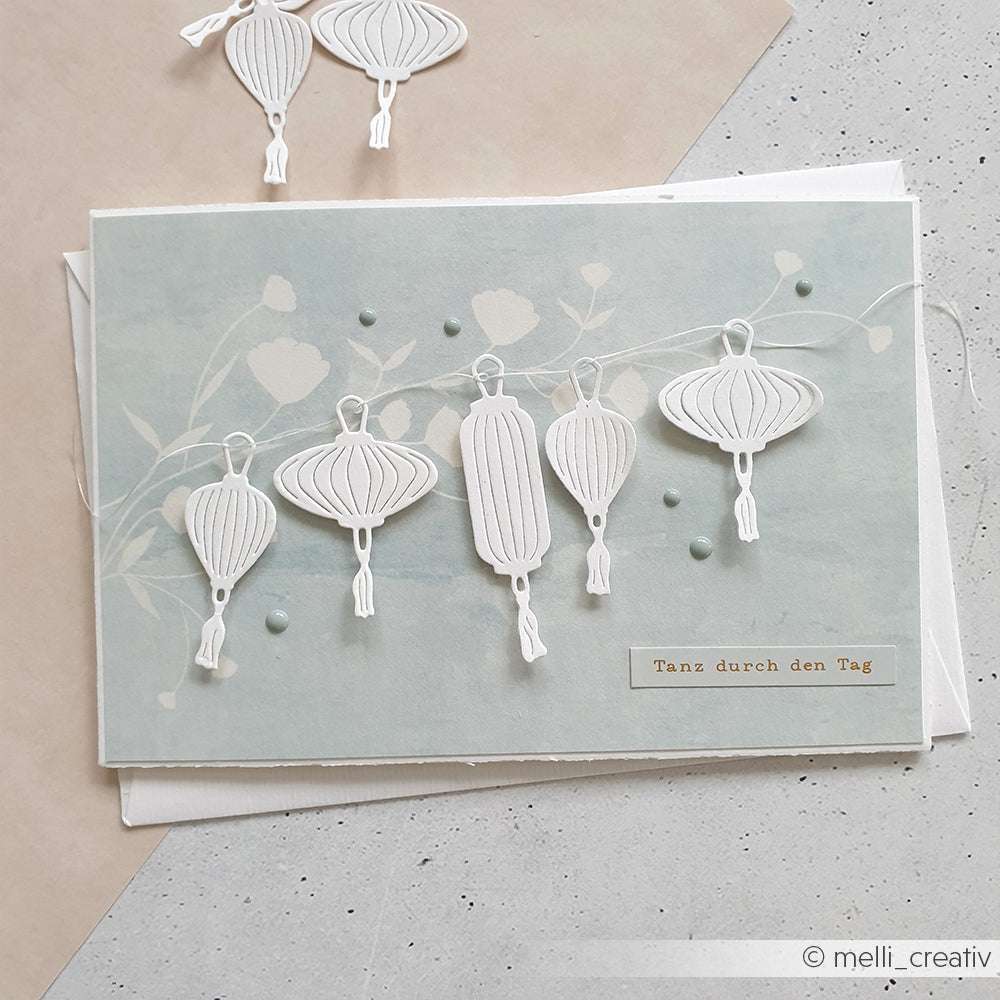 Handgemachte Karte mit weißen Papierlaternen, hergestellt mit dem Alexandra Renke PAPPcut®ies Motivstanze Lampionset, und deutschem Text "Tanz durch den Tag" auf hellblauem Hintergrund.