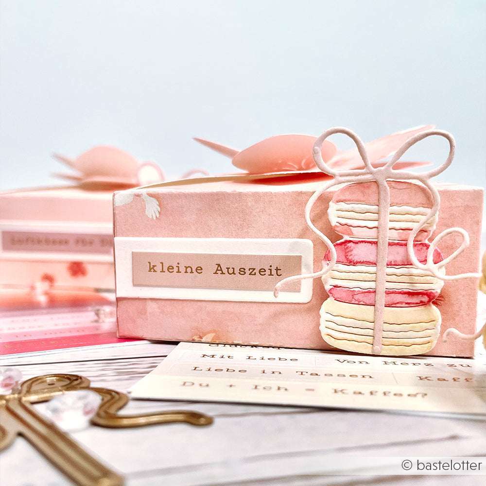 Rosafarbene Geschenkbox von Alexandra Renke mit dem Etikett "Kleine Auszeit" und einer Macaron-Illustration, umgeben von Karten und Dekorationen, die mit Deep Edge Motivstanzen hergestellt wurden, die für charmante Details sorgen.