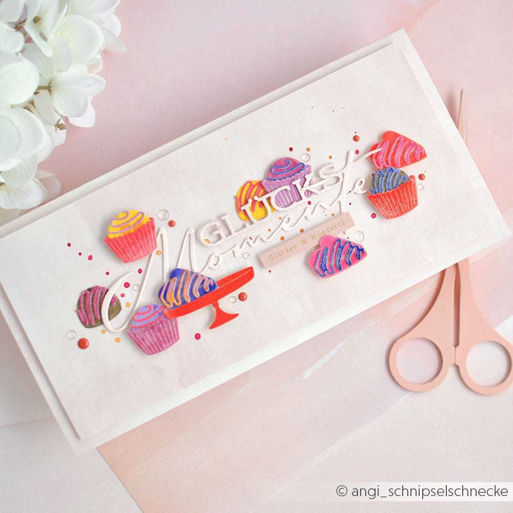 Handgemachte Karte mit bunten Cupcakes aus Alexandra Renke PAPPcut®ies und dem Text "Glücksmomente", mit rosa Schere und Blumen im Hintergrund.
